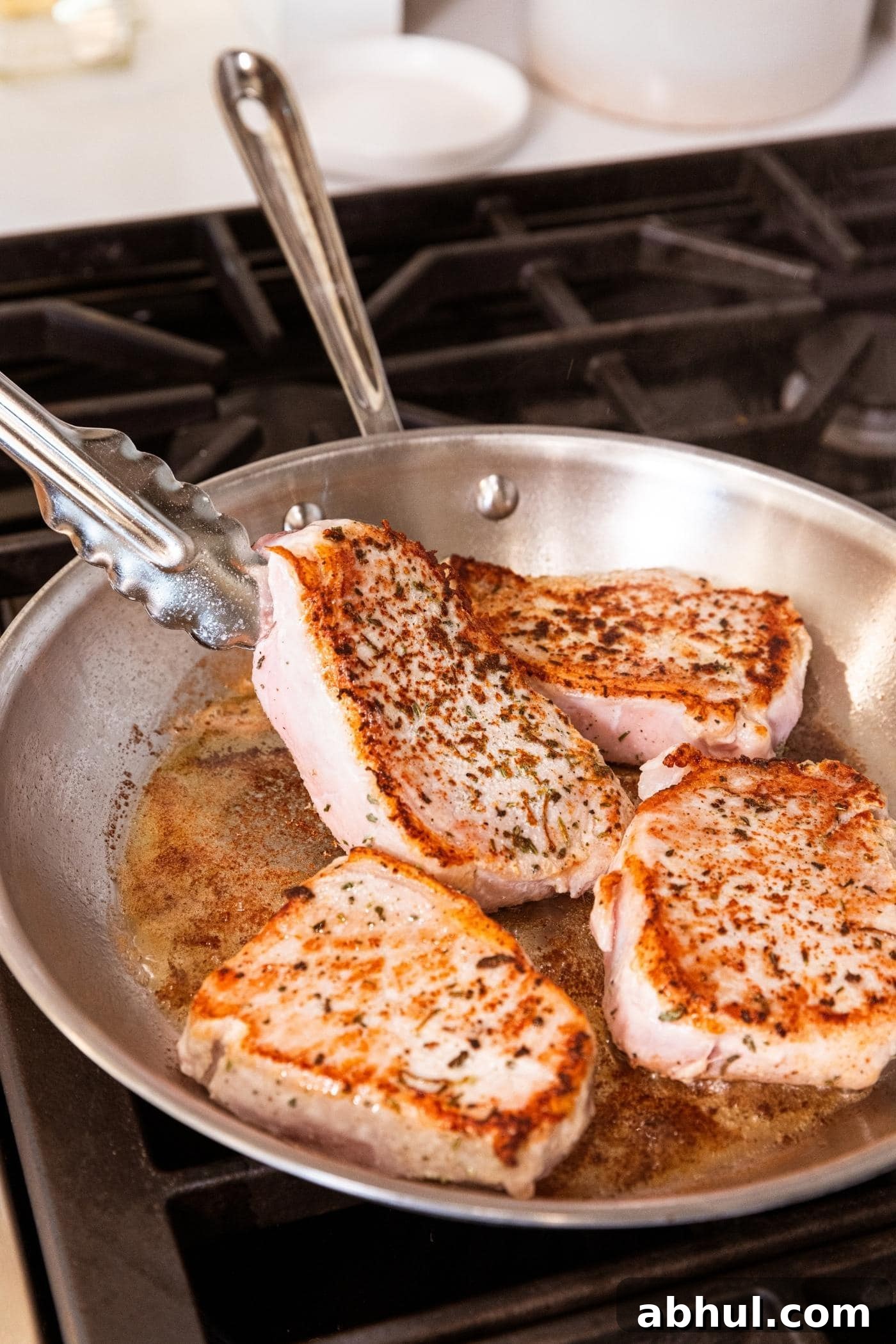 Tongs turning browned pork chop in metal skillet on stovetop with pan juices collected in pan.
