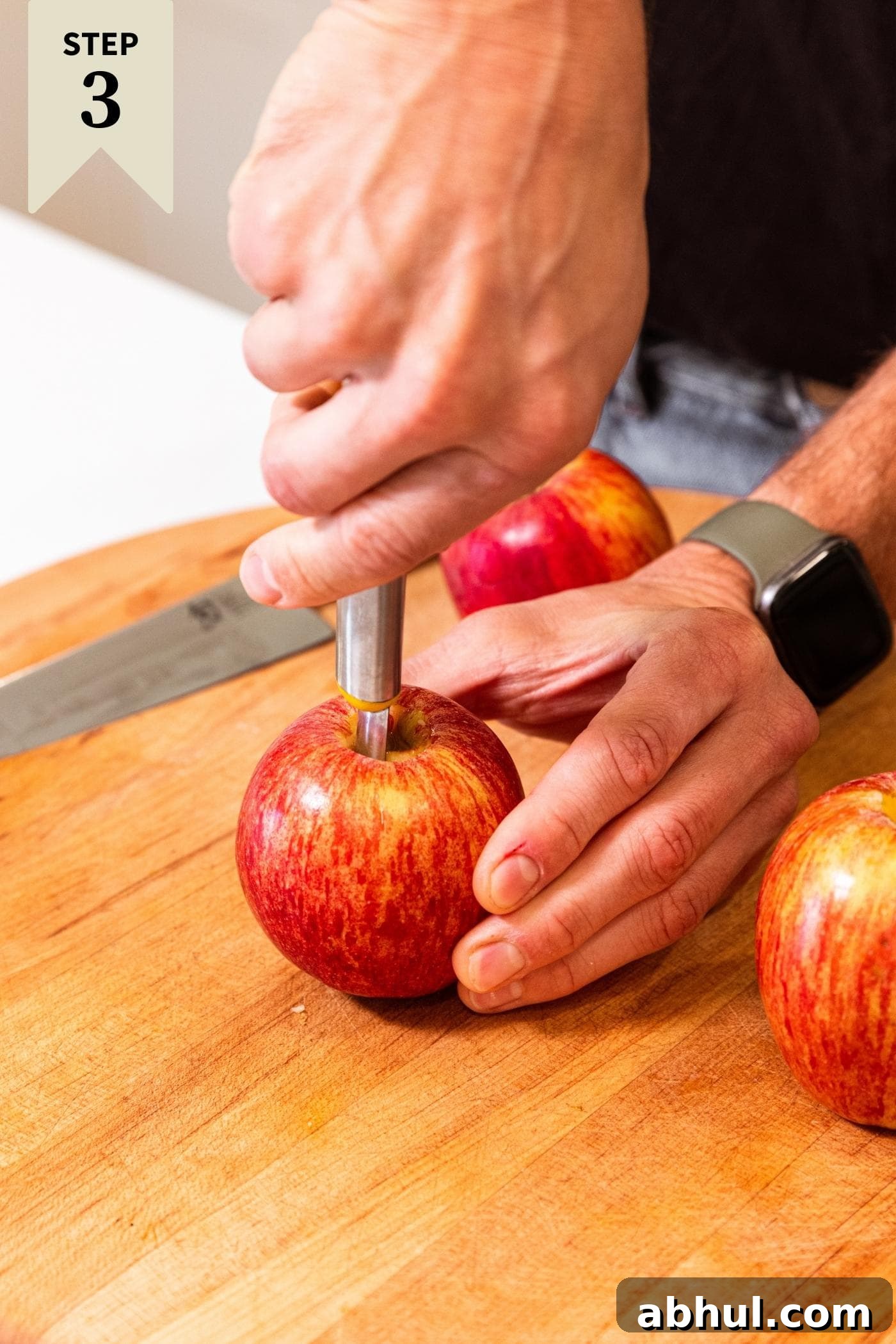 Two hands holding an apple using tool to core out the center of the apple with more apples sitting around waiting as well.
