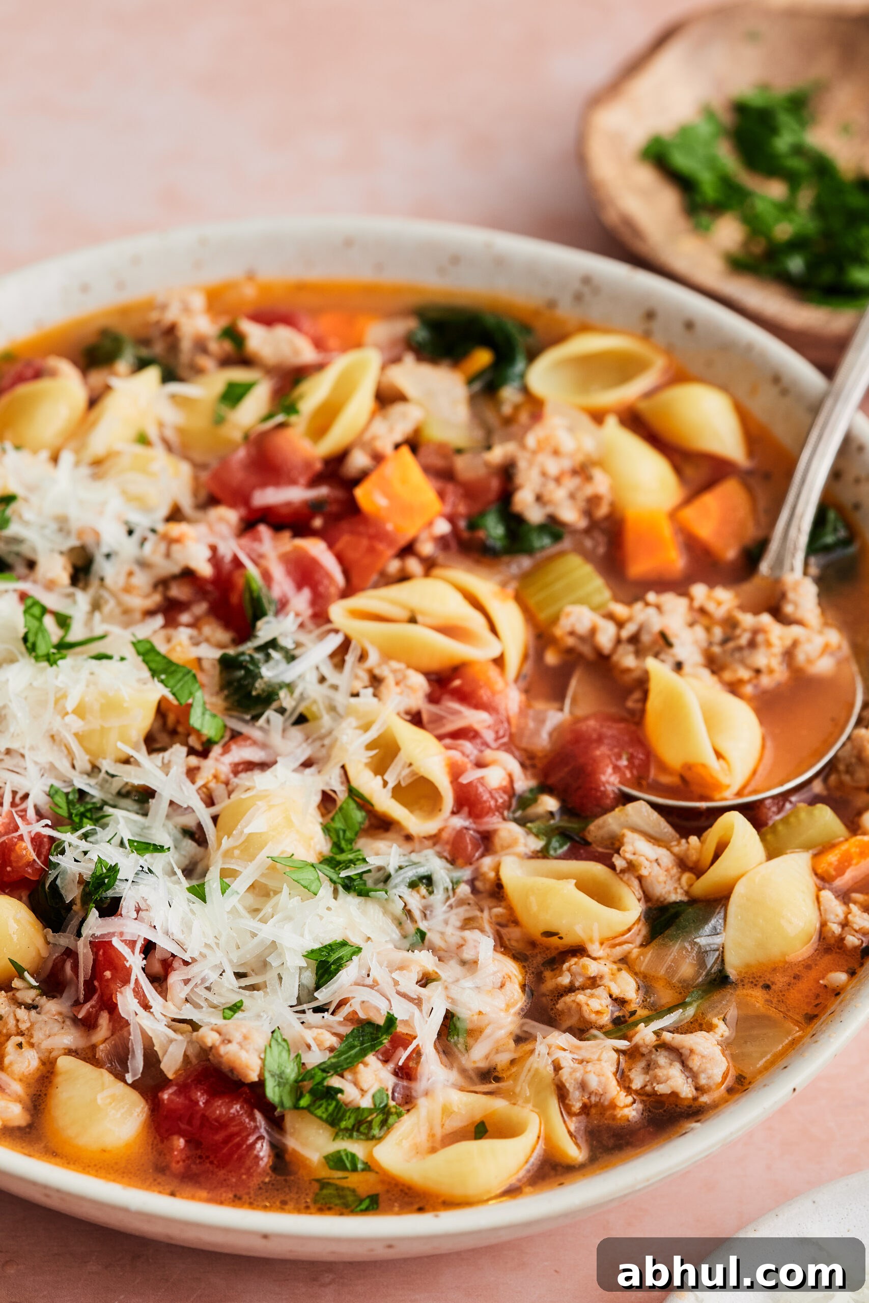 Bowl of Italian sausage soup topped with cheese and fresh herbs, with a spoon scooping out a serving against a light pink background.