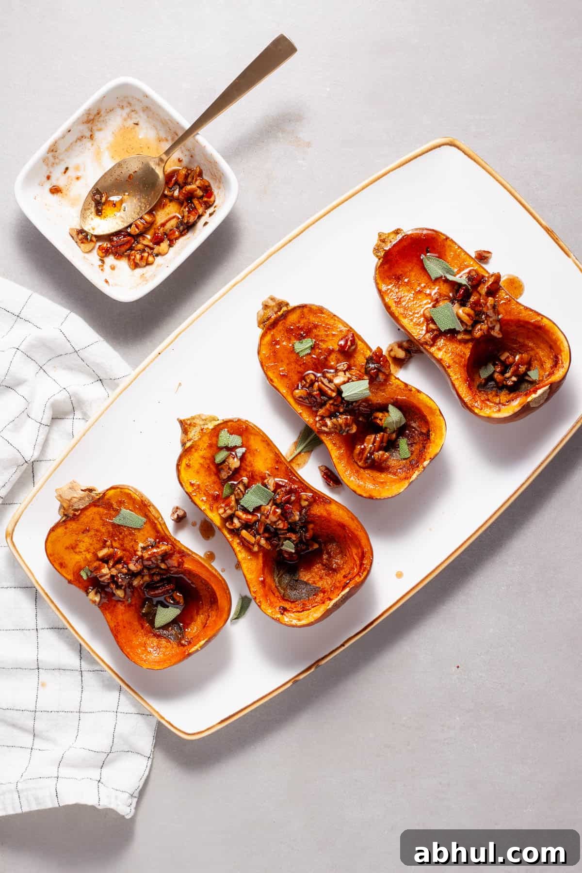 Maple-Pecan Roasted Honeynut Squash Recipe 11 Roasted honeynut squash topped with candied pecans and sage on a white plate with a platter of more off center.