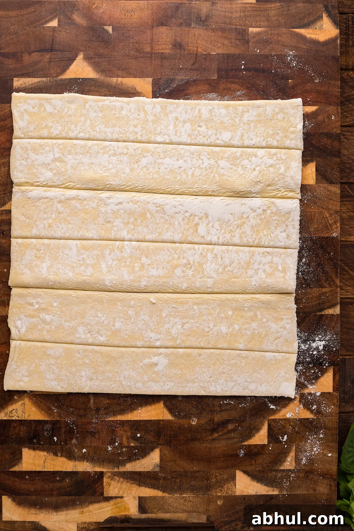 An unrolled sheet of raw, floured puff pastry lies on a wooden surface.