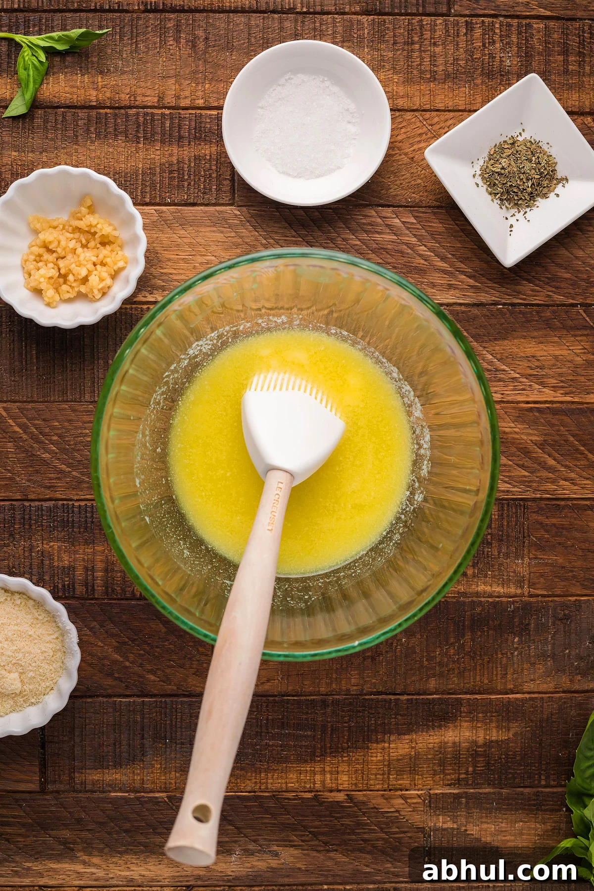 A glass bowl with melted butter and a brush sits next to dishes of minced garlic, salt, herbs, and grated cheese.