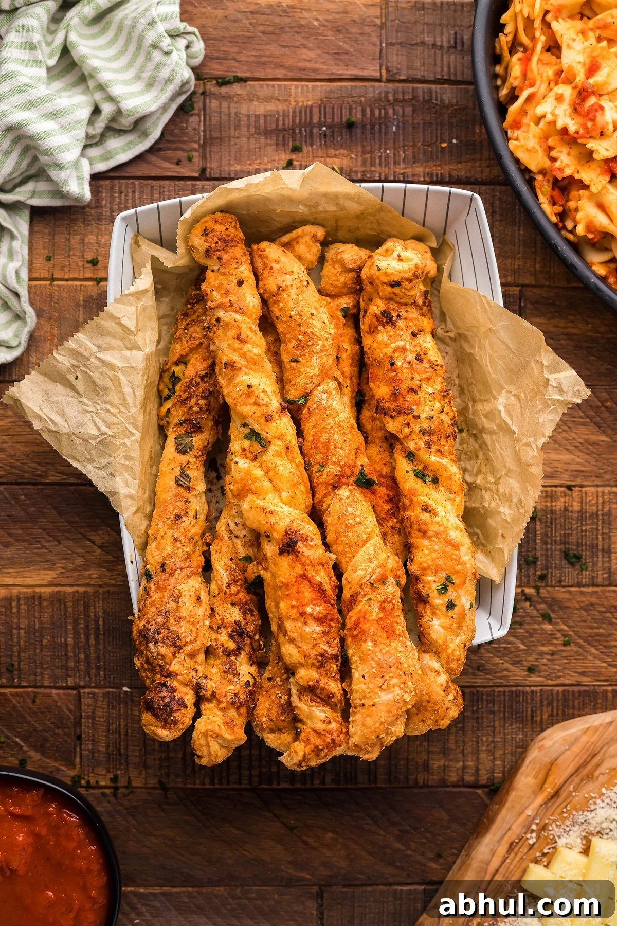 A basket of delectable Air Fryer Puff Pastry Cheese Twists sits on parchment paper atop a wooden table, accompanied by a striped cloth and a nearby pasta dish—an ideal air fryer appetizer or side dish to elevate any meal.