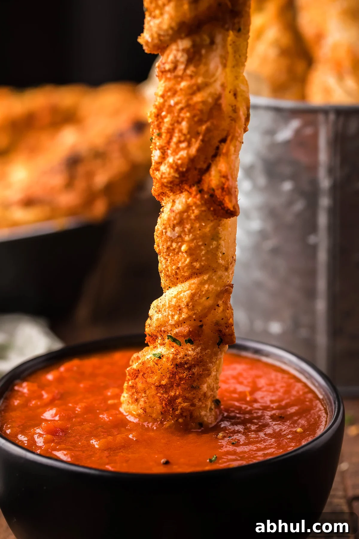 A puff pastry twist coated in seasoning, topped with parmesan and mozzarella, is being dipped into a vibrant red marinara sauce in a sleek black bowl.