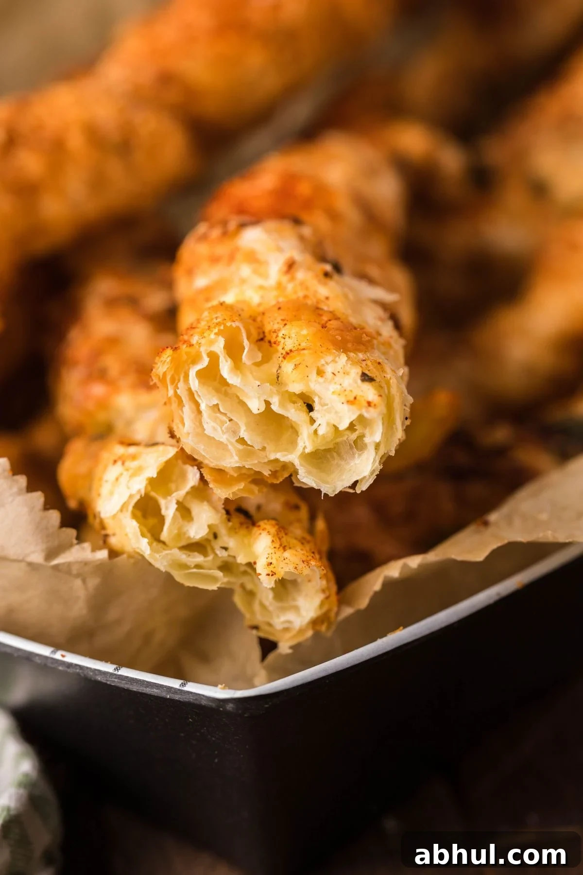 A close-up of crispy, golden-brown cheese puff pastry twists from scratch on parchment paper reveals a flaky, layered interior.