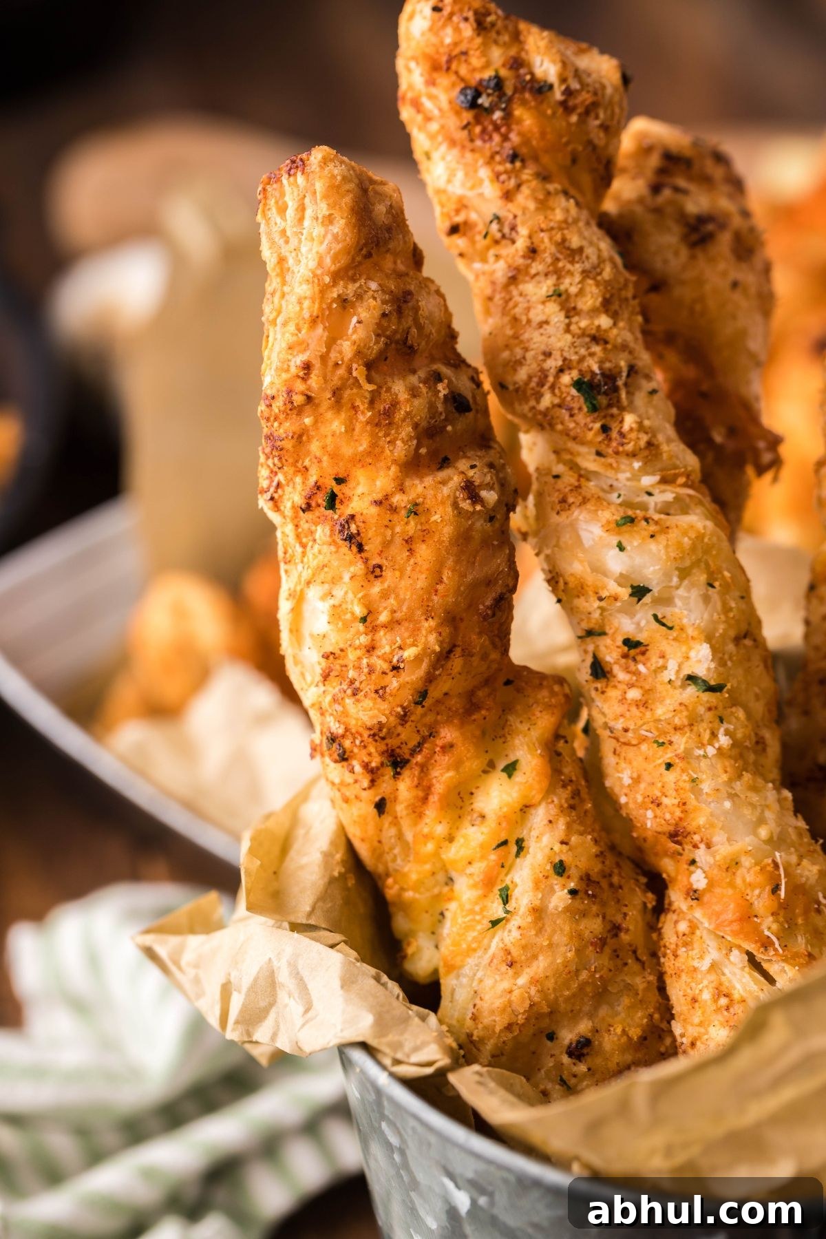 Close-up of crispy, twisted puff pastry breadsticks in a metal container lined with parchment paper, sprinkled with herbs and cheese.