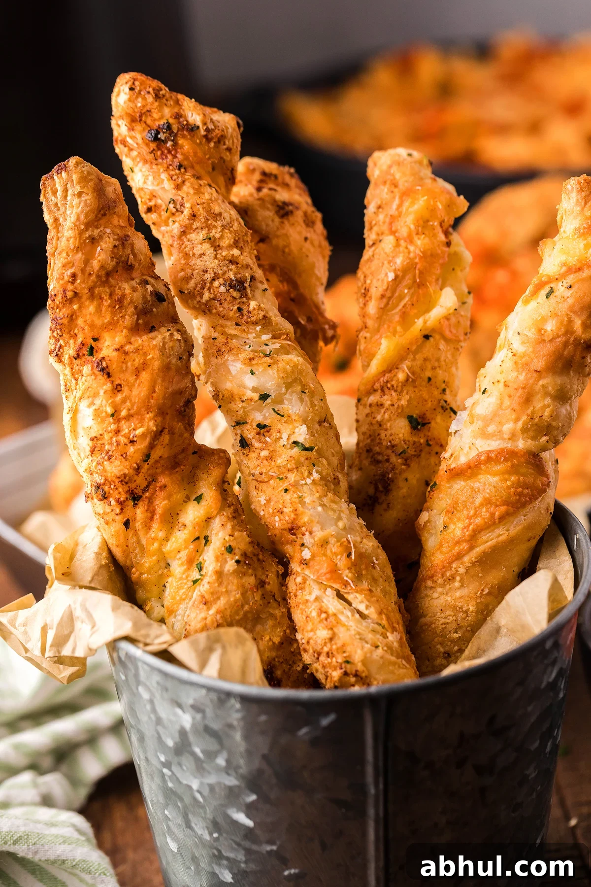 Four twisted puff pastry cheese straws with Parmesan and mozzarella sit elegantly in a metal container, neatly arranged with a napkin.