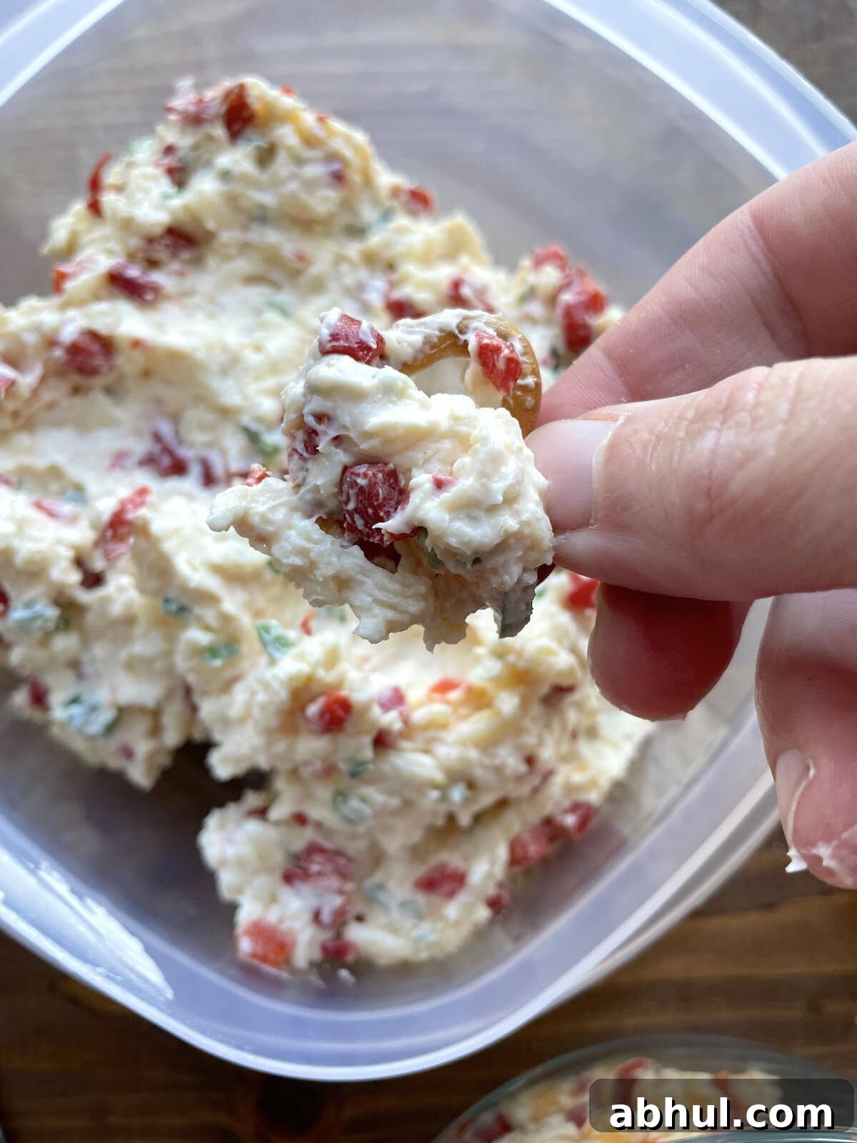 A hand holding scoop of pimento cheese with jalapeños on a pretzel over a Tupperware.