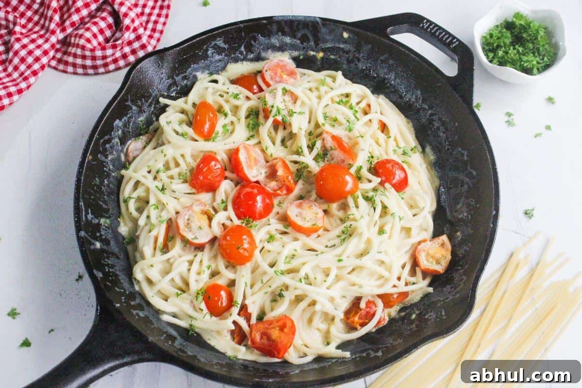 A skillet filled with creamy spaghetti topped with halved cherry tomatoes and chopped herbs, with uncooked pasta and parsley nearby on a white surface.