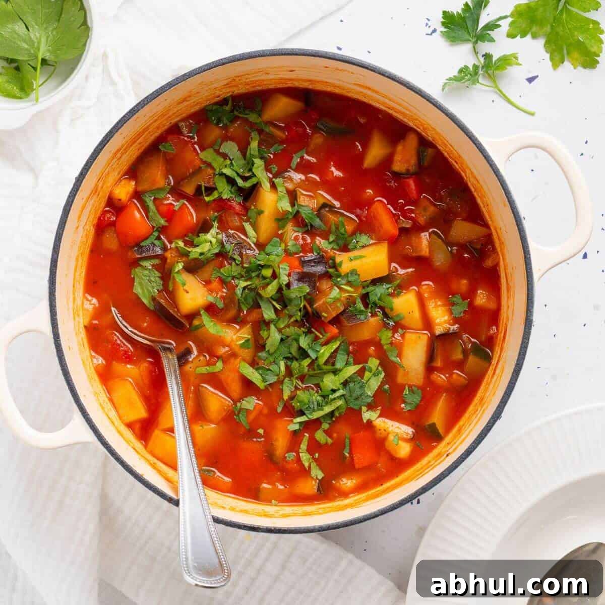 A vibrant ciambotta vegetable stew in a white pot, garnished with chopped herbs, with a spoon inside, served on a cloth-covered table.