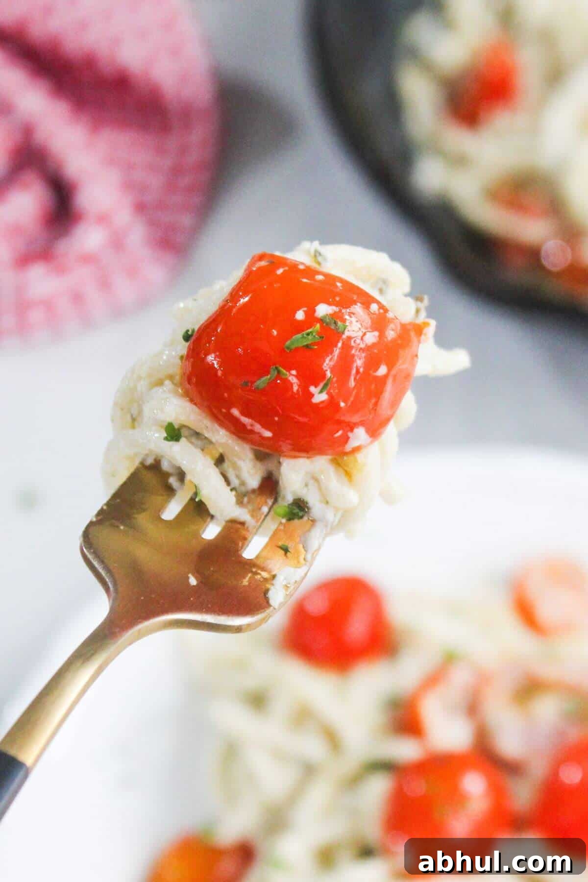 A close-up of a fork holding spaghetti and a roasted cherry tomato, with more pasta and tomatoes visible on a plate in the background.