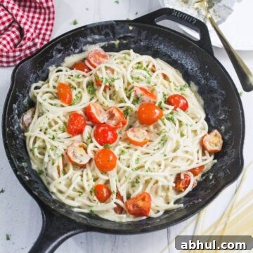 A cast iron skillet filled with spaghetti in a creamy white sauce, garnished with cherry tomatoes and chopped herbs.