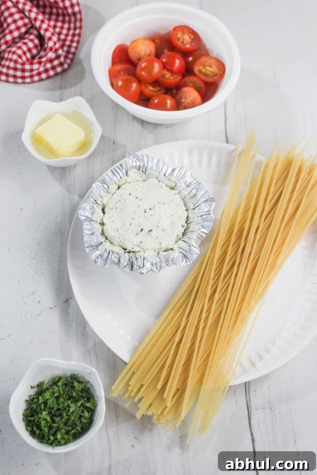 Uncooked spaghetti, a foil-wrapped cheese round, halved cherry tomatoes, butter, and chopped herbs are arranged on a white plate and table.