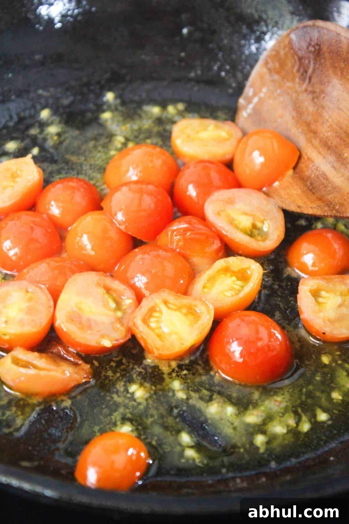 Halved cherry tomatoes cooking in a skillet with minced garlic and oil, being stirred with a wooden spoon.