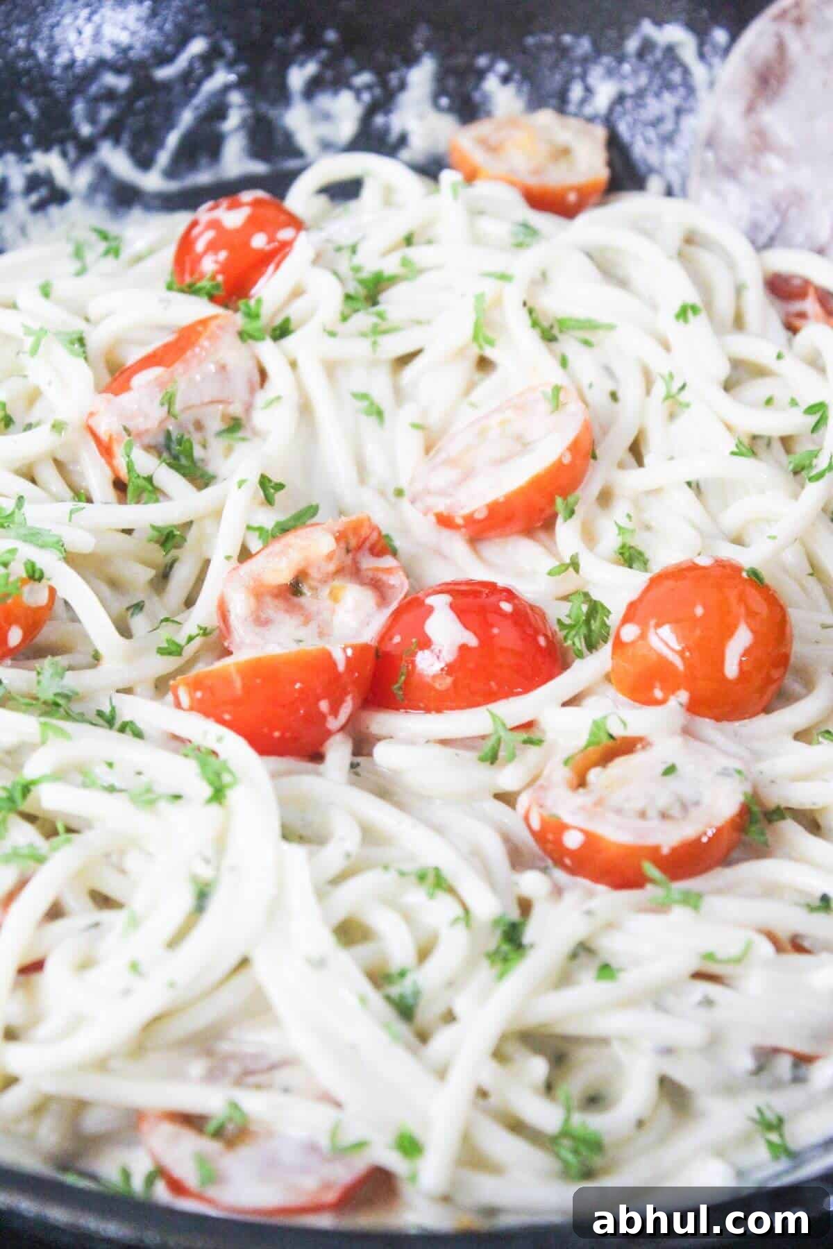 Close-up of spaghetti in a creamy white sauce with cherry tomato halves and chopped parsley, served in a pan.