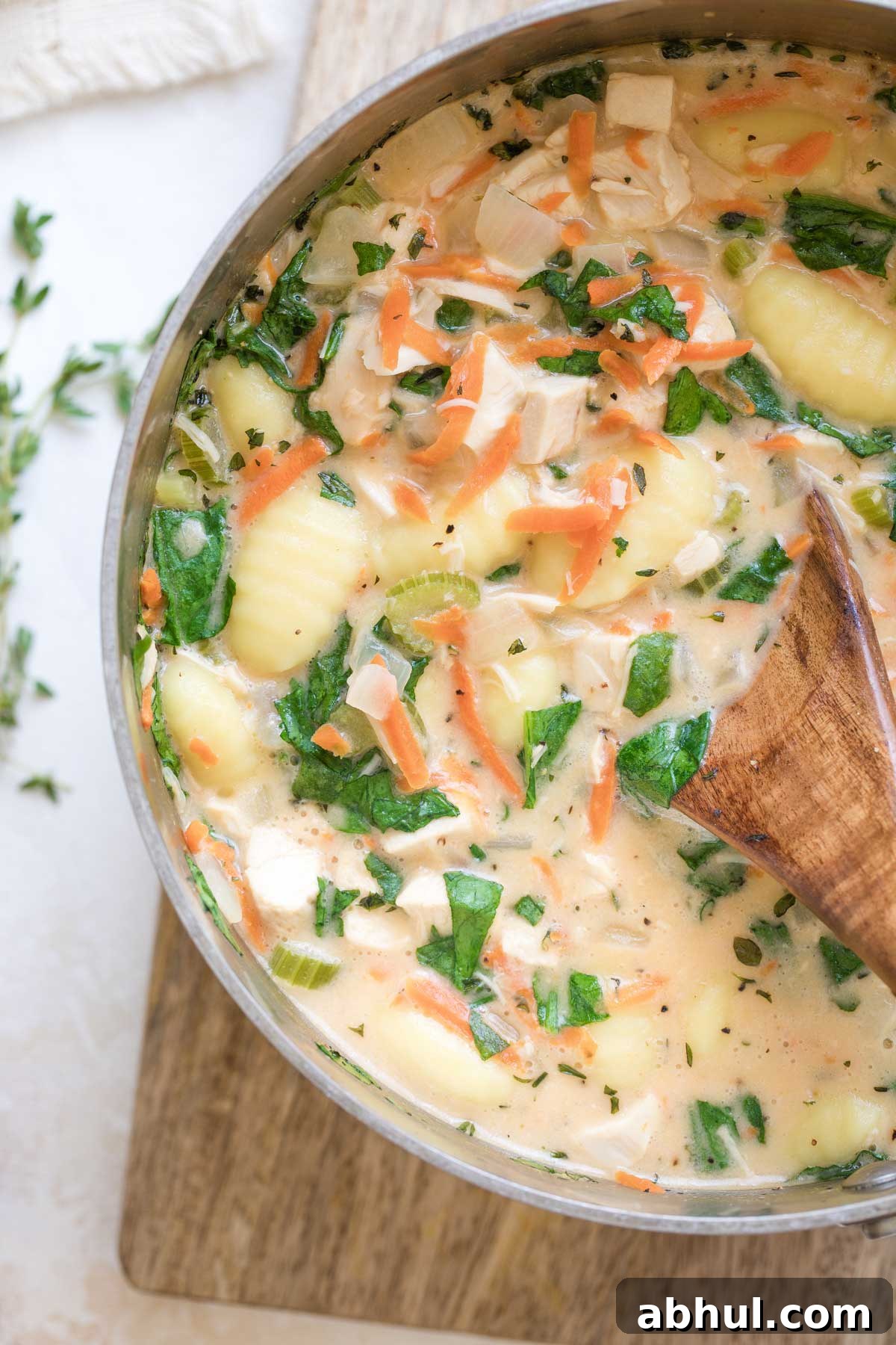 Overhead of left half of pot full of finished soup, on wooden board with spoon dipping in to help show texture of chunks of chicken and gnocchi.