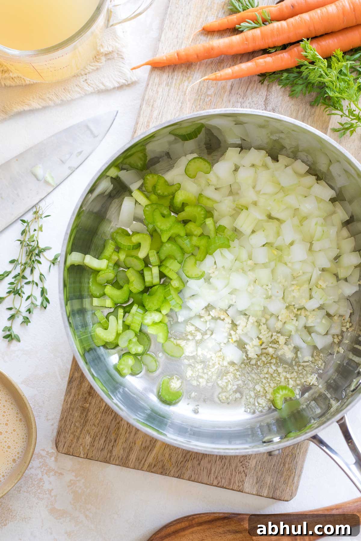 Overhead of soup pot with uncooked celery, onion and garlic added, surrounded by whole carrots, chicken broth and utensils.