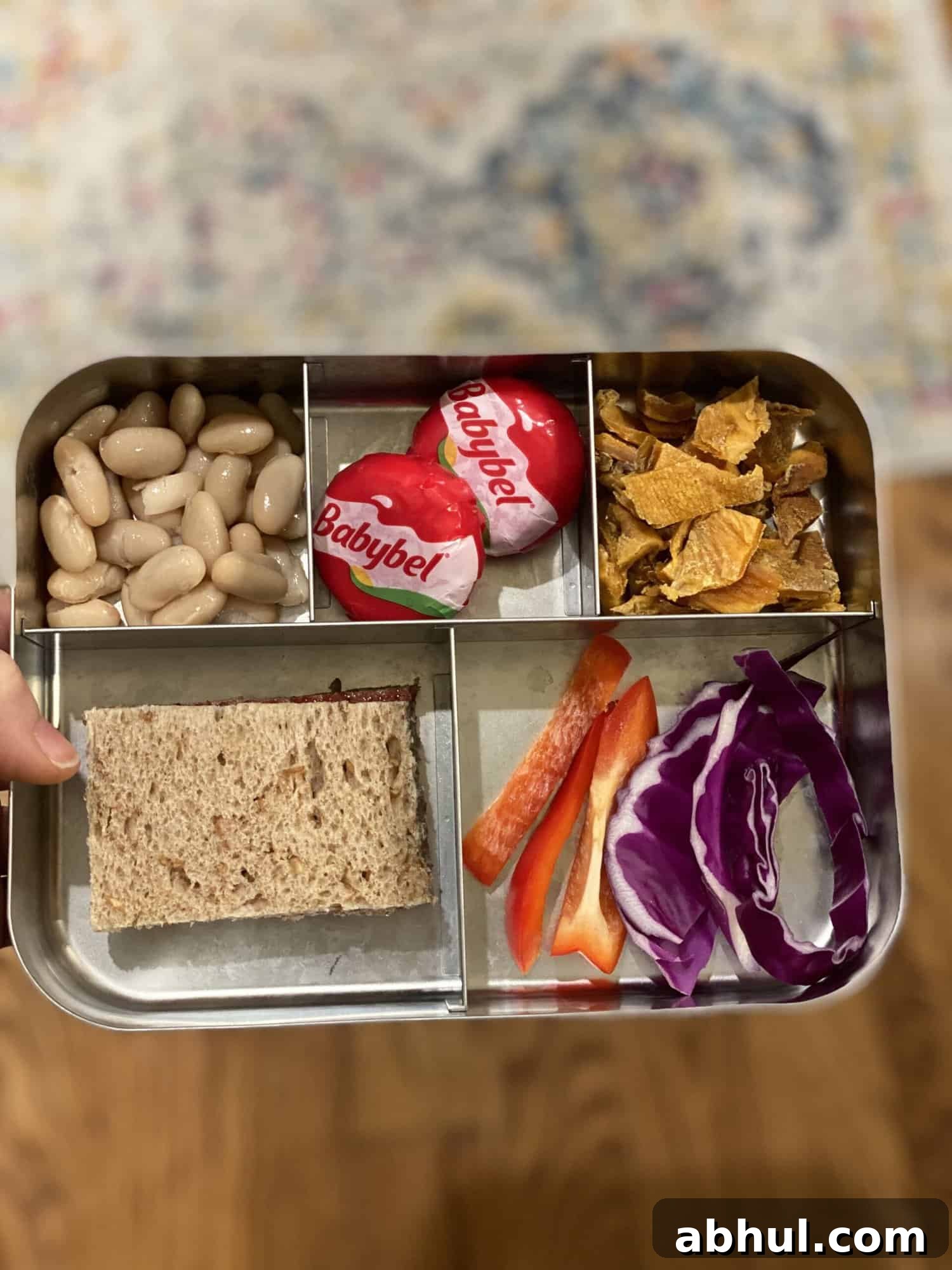 packed lunch for 2 year old with a sandwich on whole wheat bread, babybel cheese, white beans, dried mango, red pepper and cabbage