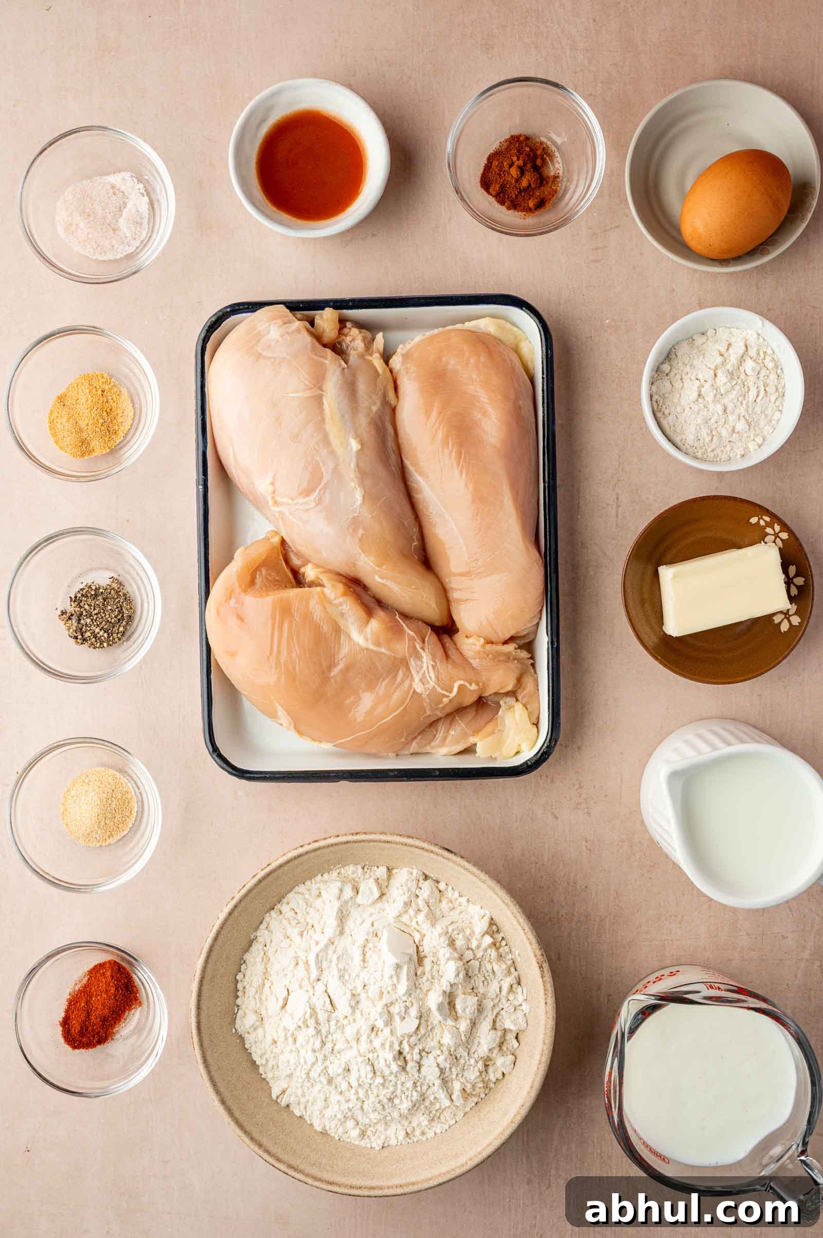 Crispy Chicken-Fried Chicken with Classic White Gravy 3 Top-down view of raw chicken breasts on a tray surrounded by small bowls containing flour, spices, butter, milk, egg, and hot sauce on a light surface.
