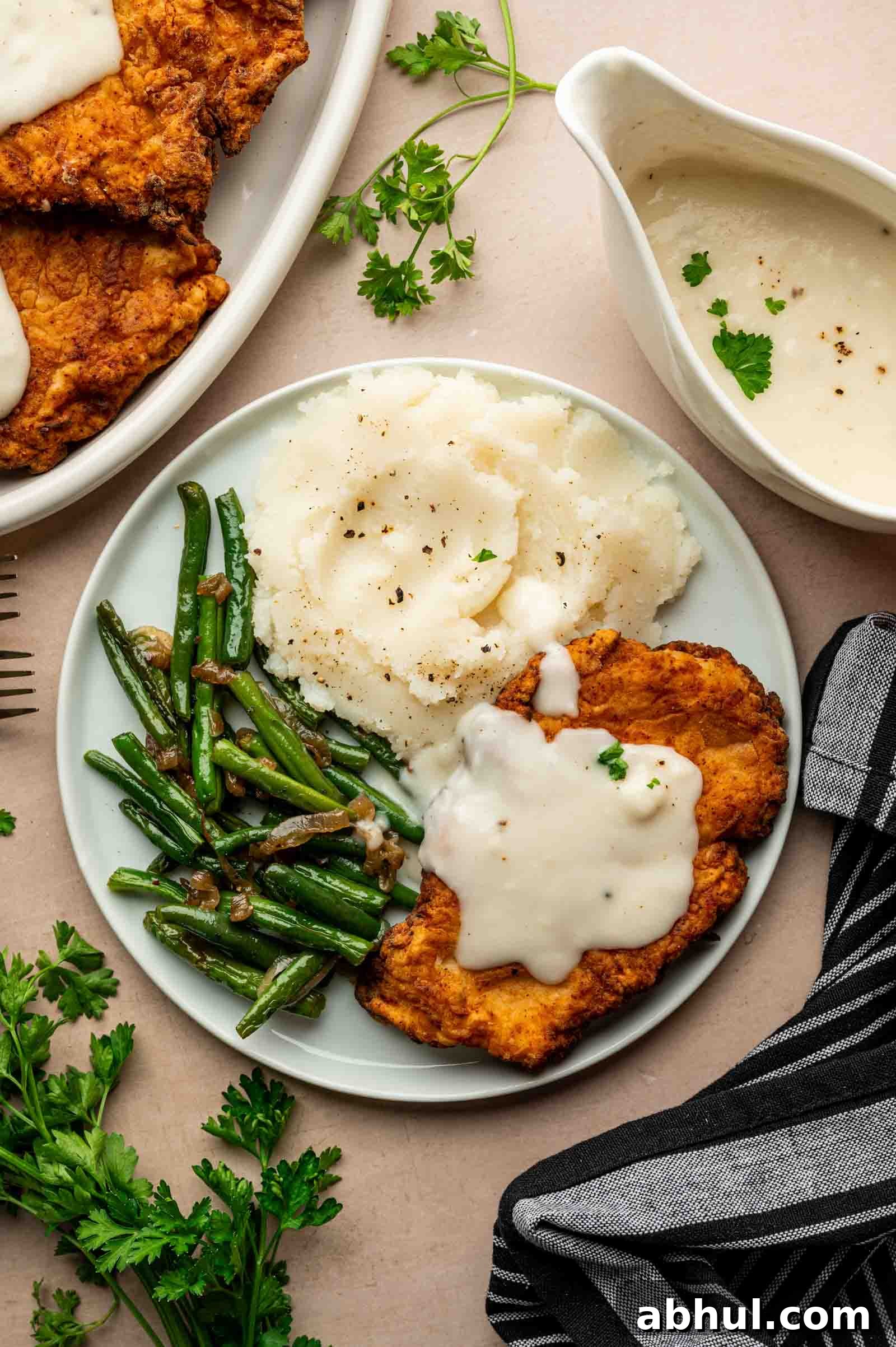 Crispy Chicken-Fried Chicken with Classic White Gravy 4 A plate with chicken fried chicken topped with white gravy, mashed potatoes, and sautéed green beans, served beside a bowl of extra gravy and fresh herbs.