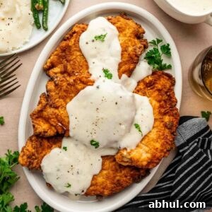 Crispy Chicken-Fried Chicken with Classic White Gravy 6 Plate of chicken fried chicken topped with white gravy, garnished with parsley, next to a striped napkin and sides of green beans and mashed potatoes.