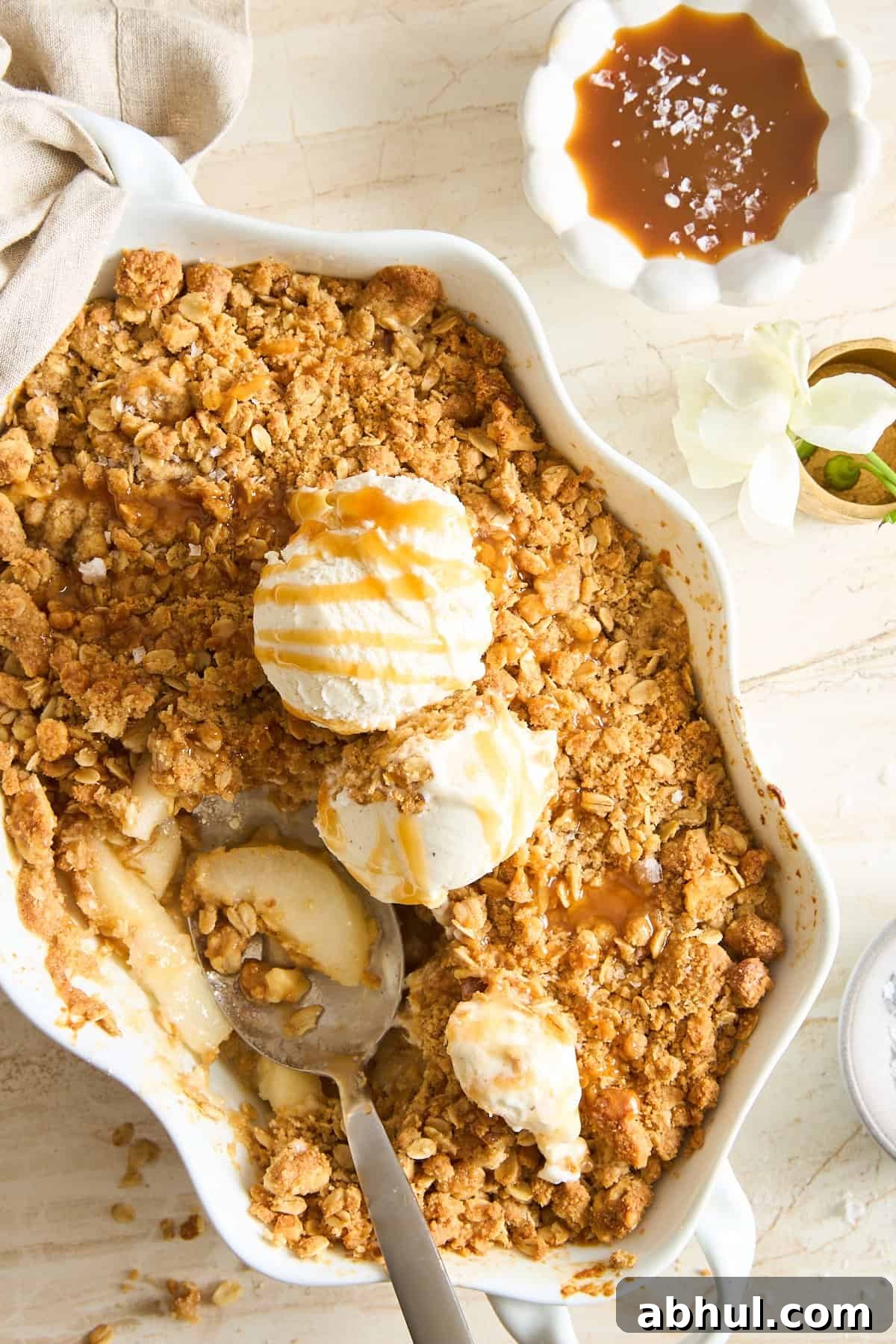 A baking dish filled with apple crisp topped with three scoops of vanilla ice cream and caramel drizzle. A spoon rests inside, and a bowl of caramel sauce sits nearby on a light-colored table.