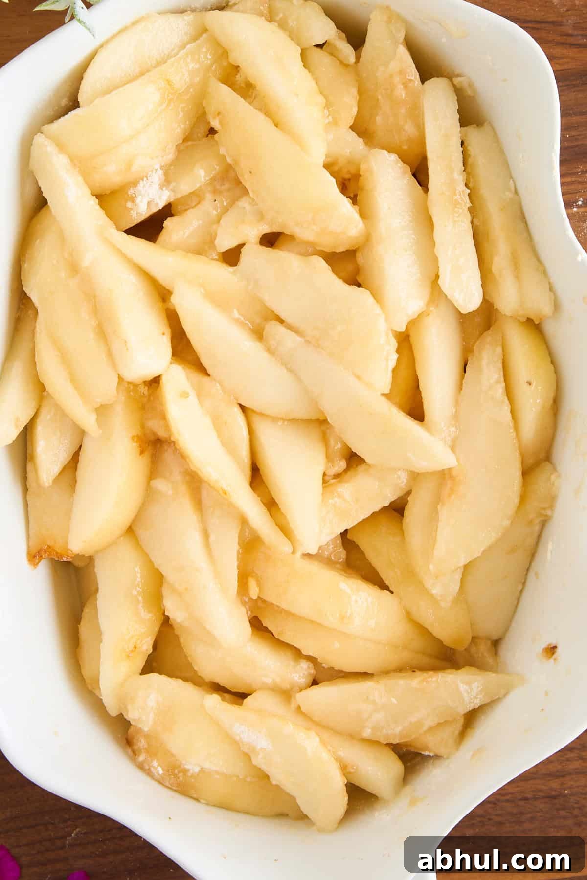 Sliced fresh pears coated with sugar and arranged in a white ceramic baking dish, ready for baking. The pears have a glossy appearance and are evenly distributed in the dish.