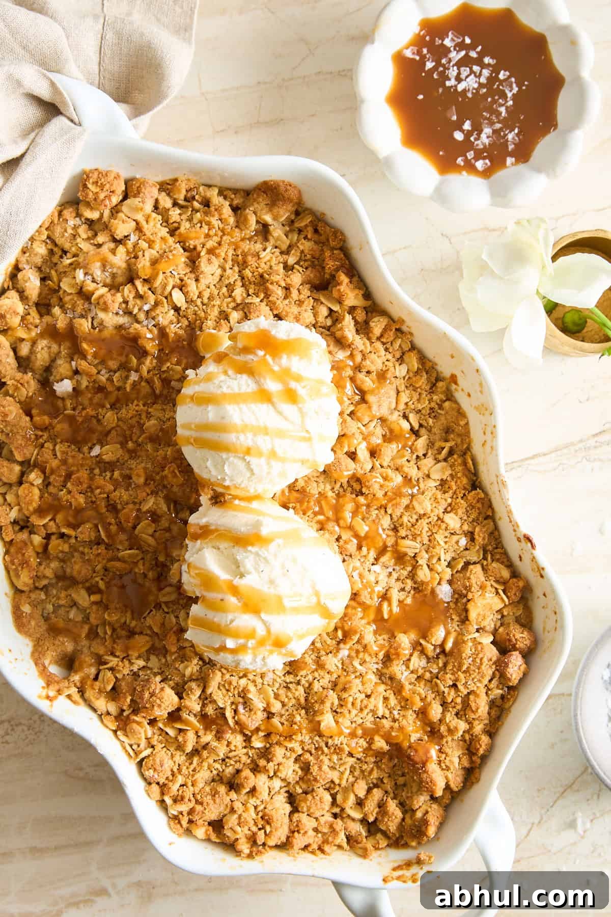 A baked apple crisp topped with two scoops of vanilla ice cream and caramel sauce in a white dish. Next to it is a bowl of caramel sauce sprinkled with sea salt and a white flower on a marble surface.