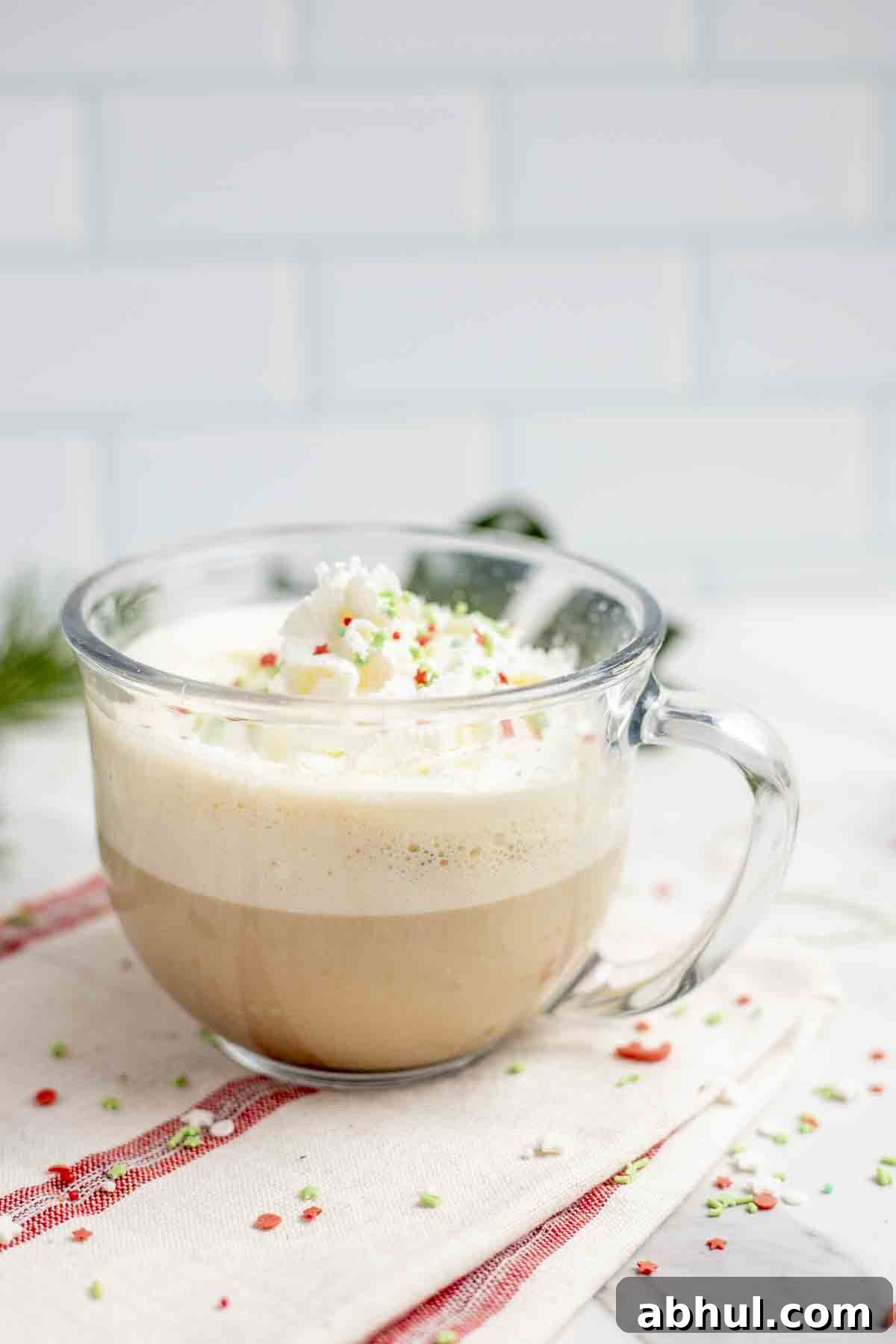 side view of a glass mug full of a eggnog latte topped with whipped cream and Christmas sprinkles. The mug sits on a white and red stripped towel with sprinkles scattered around.