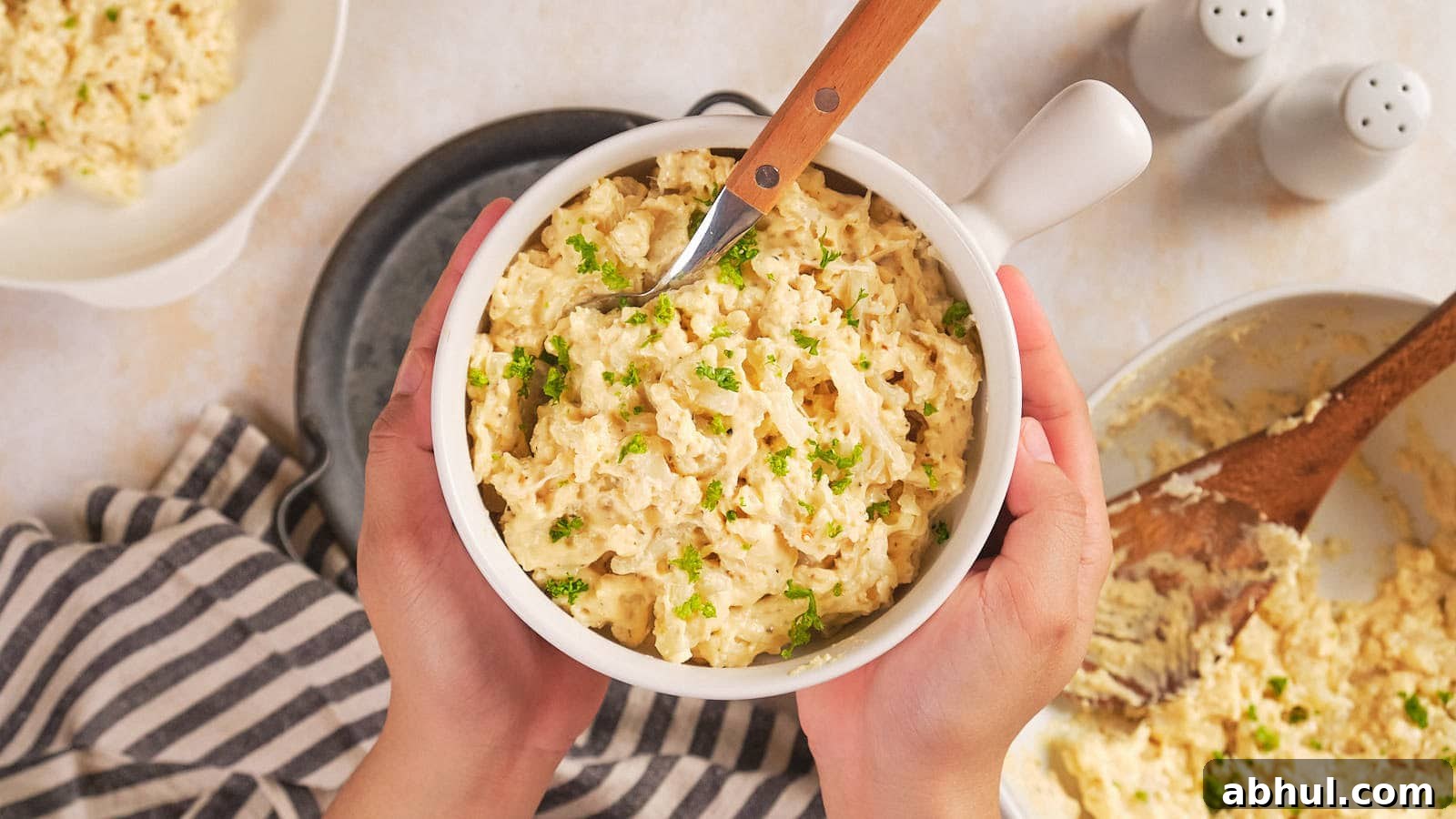 Hands holding a white bowl of cauliflower mac and cheese garnished with chopped herbs.