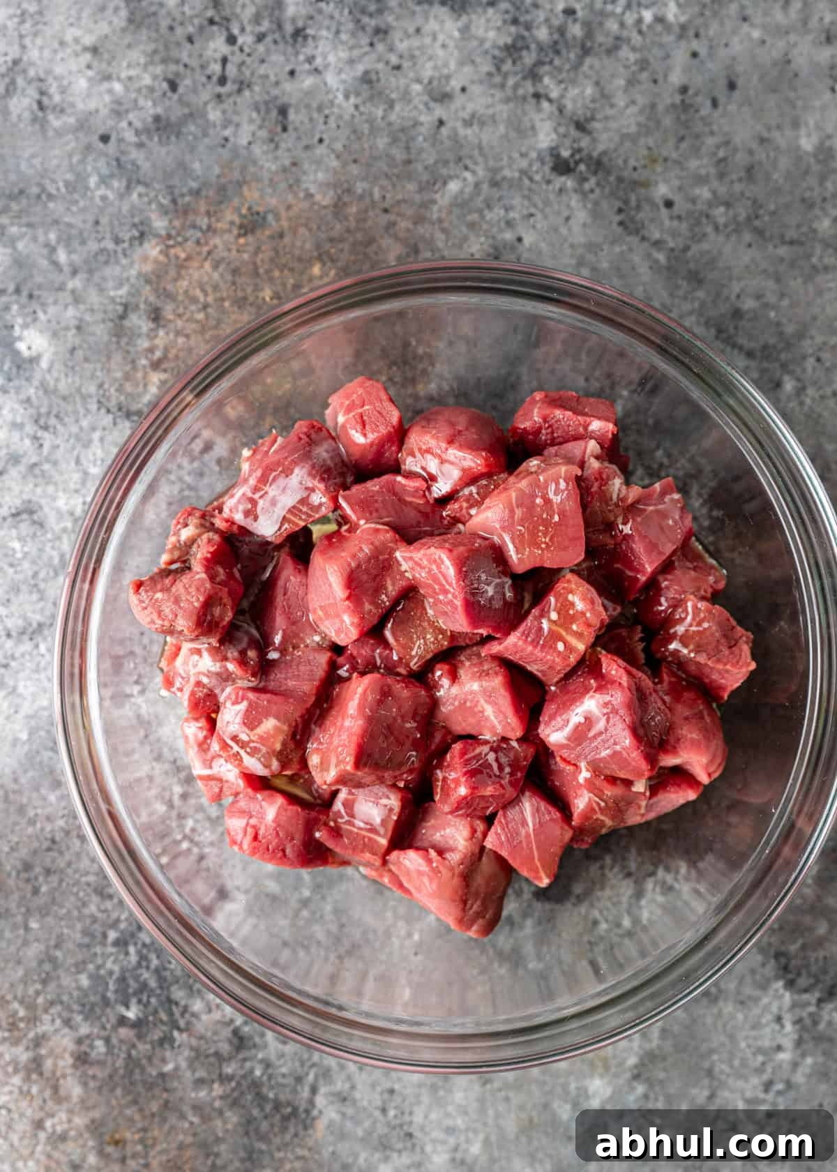 overhead process shot: cubed raw beef in a glass bowl tossed with melted butter