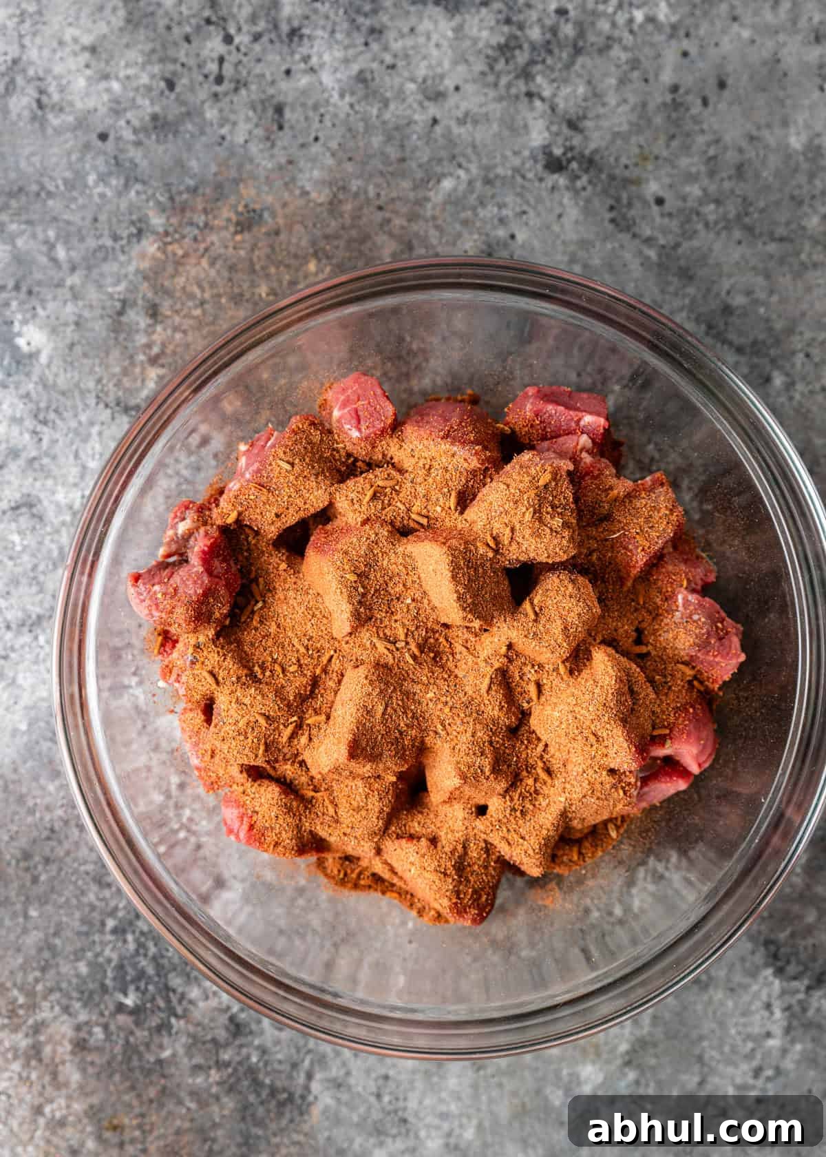 overhead process shot: cubed raw beef in a glass bowl tossed with melted butter with blackened steak seasoning on top