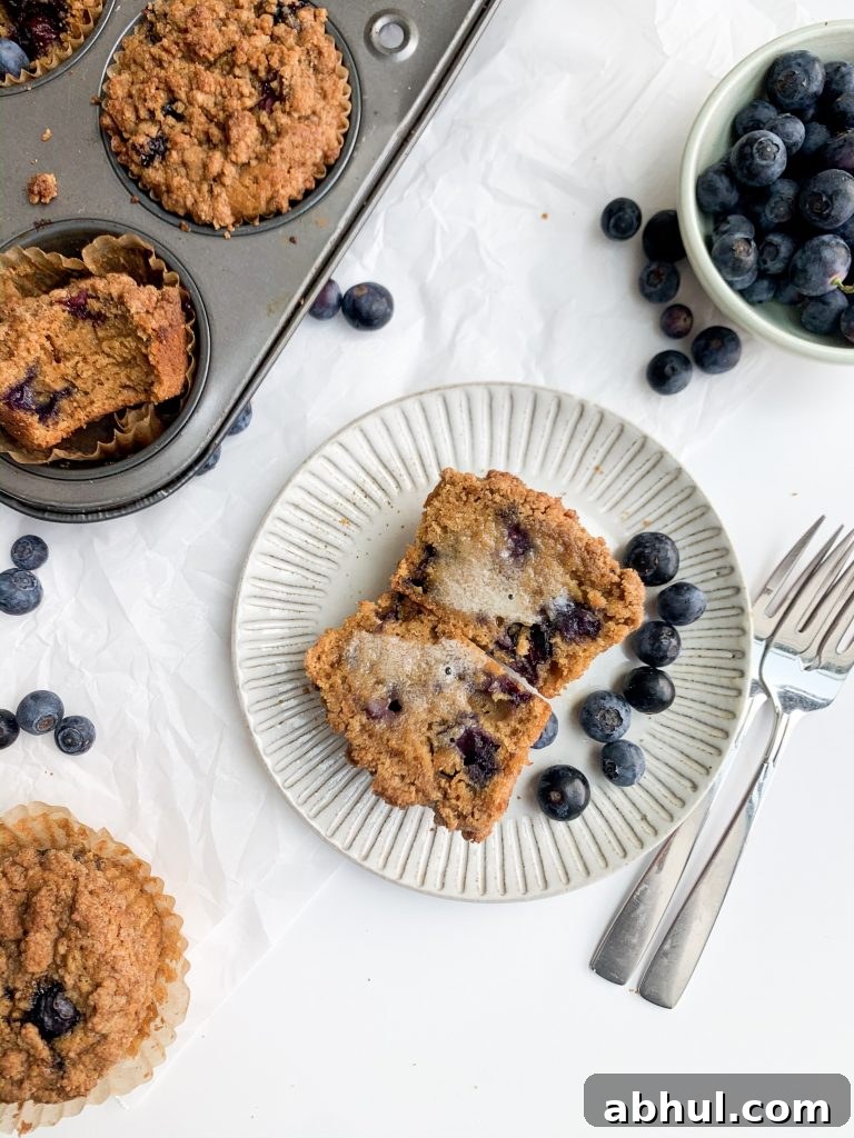 a blueberry muffin on a gray plate, a bowl of blueberries, and a muffin tin on a white background