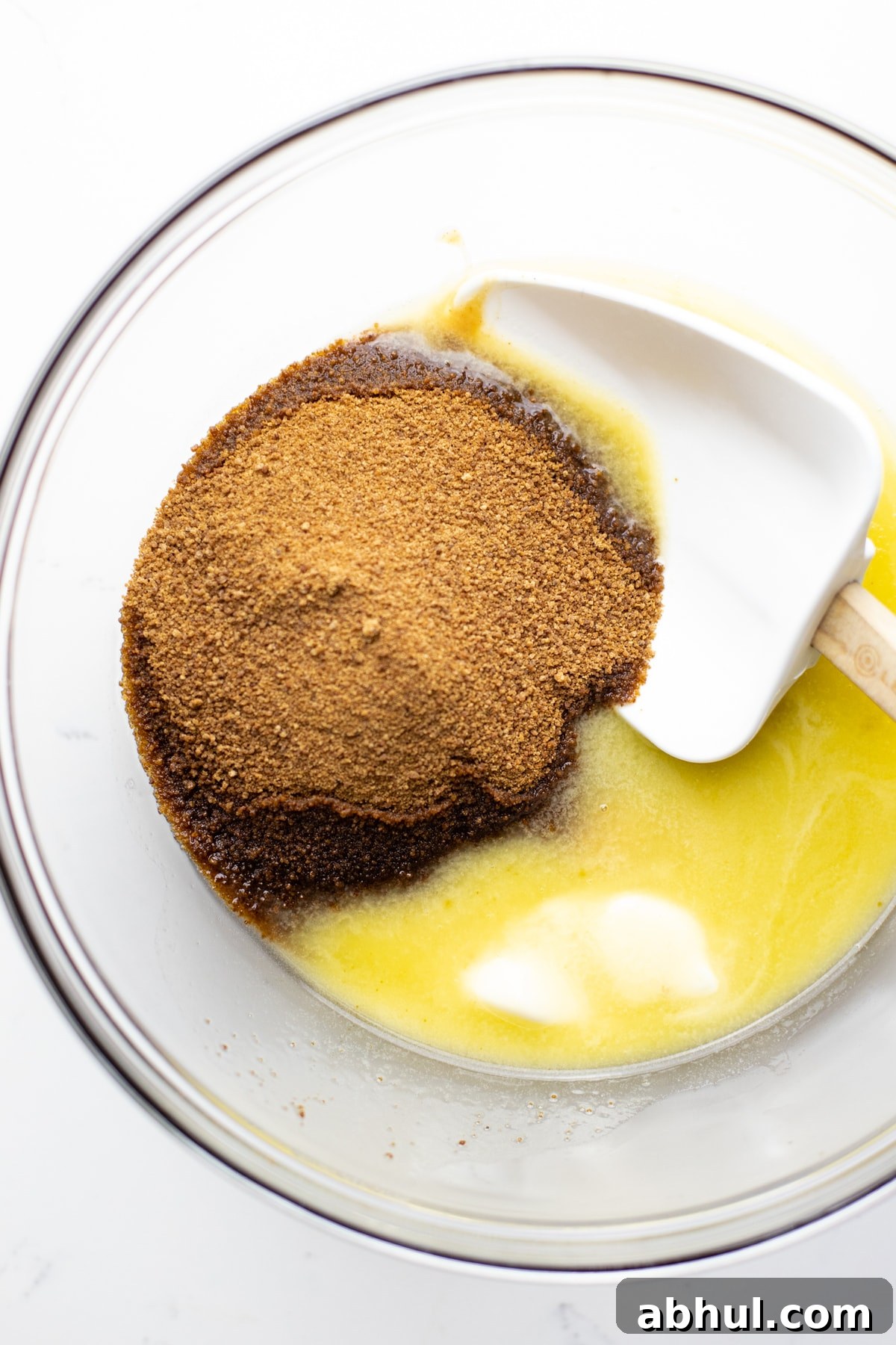 A clear bowl with coconut sugar, melted butter, and a rubber spatula.