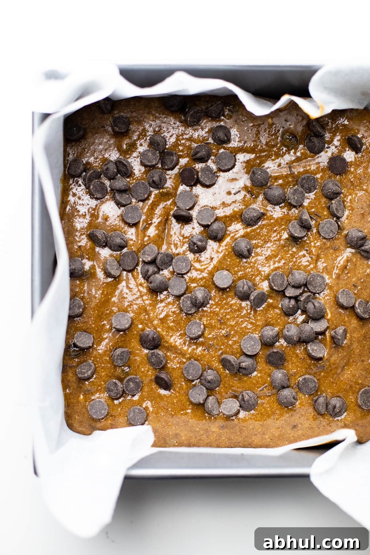 A square pan lined with parchment paper with oatmeal bar batter and chocolate chips over top.